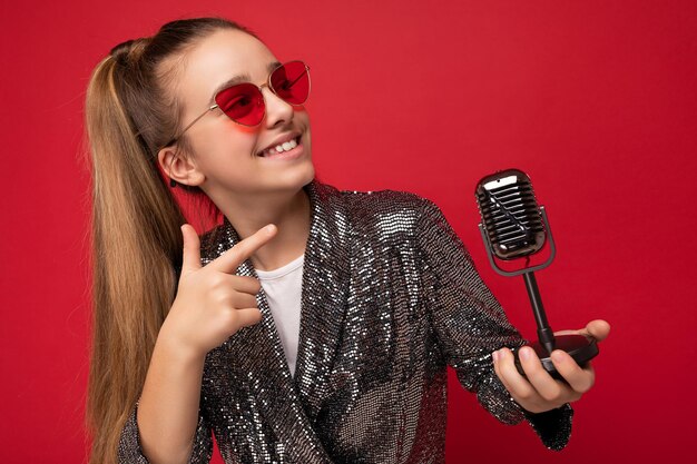 Hübscher lächelnder Brunet weiblicher Teenager, der eine stylische Glanzjacke und eine rote Brille trägt, die über Rot isoliert ist?