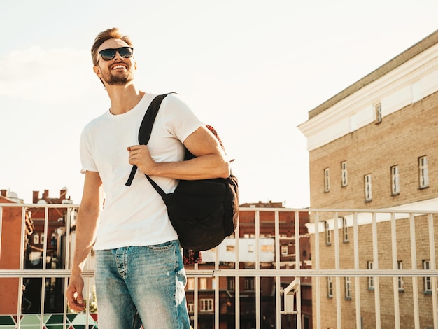 Hübscher lächelnder Hipster Lambersexuelles Modell Stilvoller Mann in weißem T-Shirt und Jeans Modemann posiert auf dem Straßenhintergrund in der Nähe des Zauns am Strand mit Schultasche in Sonnenbrille