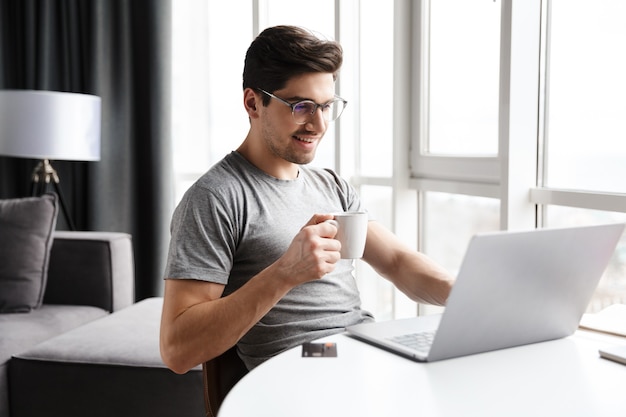 Hübscher lächelnder junger bärtiger Mann, der Freizeitkleidung mit Laptop-Computer trägt, während er zu Hause am Tisch sitzt und Kaffee trinkt