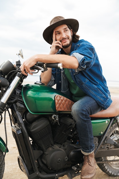 Hübscher lächelnder junger Mann mit lässigem Outfit, der auf einem Motorrad am Strand sitzt und einen Hut trägt