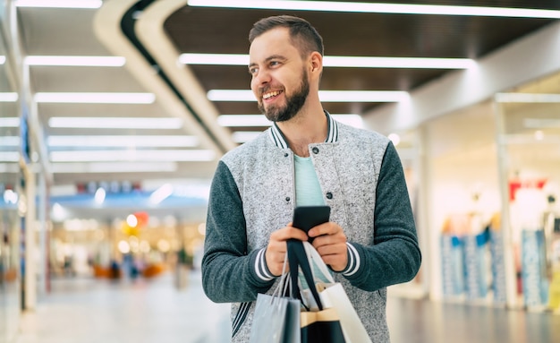 Hübscher lächelnder junger stilvoller bärtiger Mann mit Einkaufstaschen verwendet Smartphone während des Gehens in der Mall