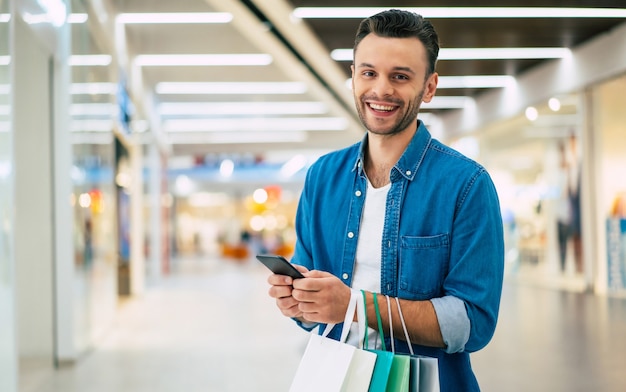 Hübscher lächelnder junger stilvoller bärtiger Mann mit Einkaufstaschen verwendet Smartphone während des Gehens in der Mall