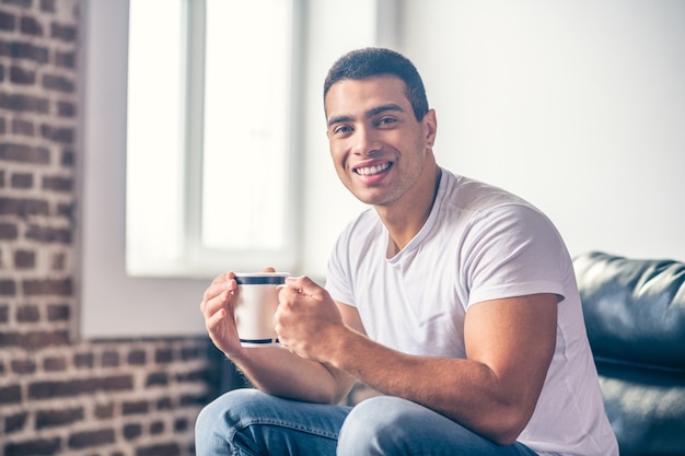 Hübscher lächelnder Kerl, der zu Hause auf einem Stuhl sitzt, mit einer Tasse Kaffee in seinen Händen.