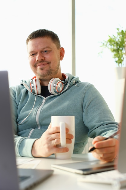 Hübscher lächelnder männlicher Student, der Online-Bildungsdienst verwendet und Tee oder Kaffee trinkt.