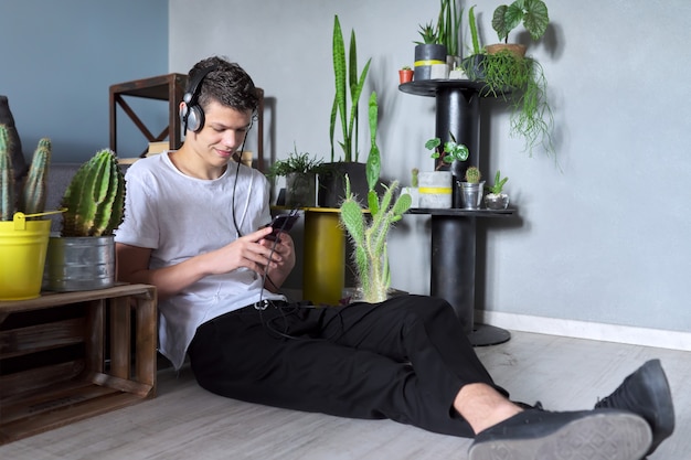 Hübscher lächelnder Teenagerjunge in Kopfhörern mit Smartphone, der zu Hause sitzt, Musik-Audio-Blogbuch hört, sich ausruht, spielt, studiert. Bildung Freizeit Lifestyle Teenager Konzept