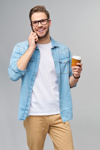 Hübscher lässiger Mann im blauen Jeanshemd, der sein Telefon und Tasse Kaffee hält, um über graue Wand des Studios zu stehen