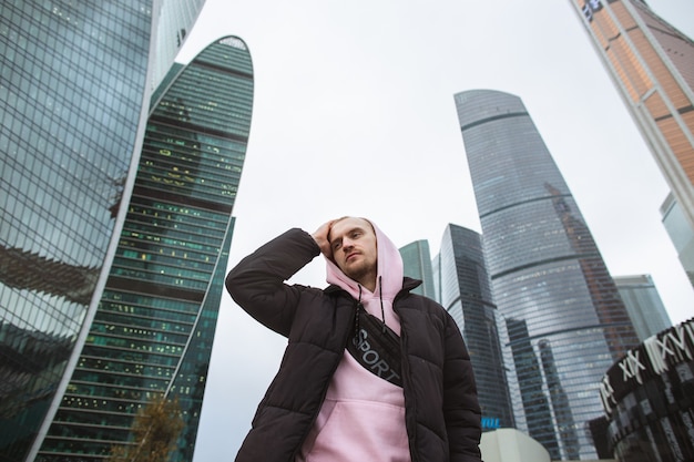 Hübscher lässiger Mann in der schwarzen Jacke und im rosa Kapuzenpulli, die auf einer Wolkenkratzeransicht stehen.