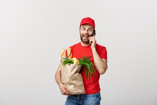 Hübscher Lebensmittelgeschäftlieferer, der mit Mobile spricht und Lebensmittelgeschäftpapiertüte hält.