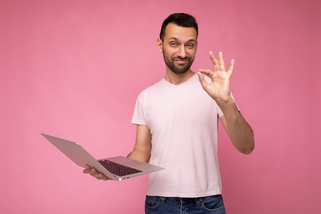 Hübscher lustiger Brunet-Mann, der Laptop-Computer hält und Geste zeigt, die Kamera im T-Shirt auf isolierter rosa Oberfläche in Ordnung betrachtet