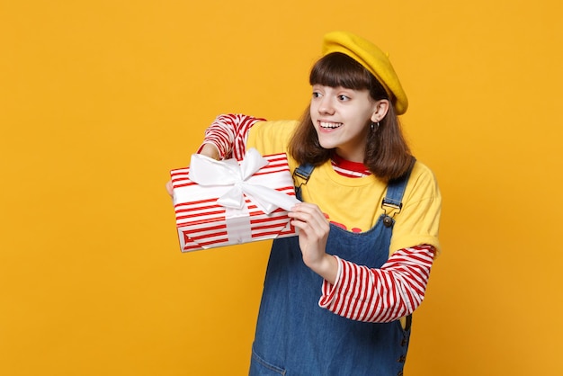 Hübscher Mädchen-Teenager in französischer Baskenmütze, Denim-Sommerkleid, das zur Seite schaut und rot gestreifte Geschenkbox mit Geschenkband isoliert auf gelbem Hintergrund hält. Menschen Emotionen, Lifestyle-Geburtstags-Urlaubskonzept.