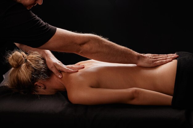 hübscher männlicher Masseur, der ein Mädchen auf schwarzem Hintergrund massiert. Konzept der therapeutischen entspannenden Massage