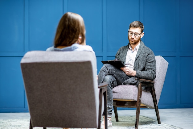 Hübscher männlicher Psychologe, der dem weiblichen Klienten zuhört, der während der psychologischen Sitzung im blauen Büroinnenraum sitzt