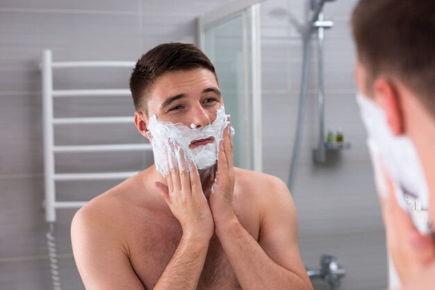 Hübscher männlicher Schmierschaum für die Rasur im Gesicht, der vor dem Spiegel im modernen gefliesten Badezimmer zu Hause steht