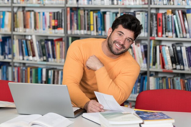 Hübscher männlicher Student mit Laptop und Büchern, die in einer High School-Bibliothek arbeiten