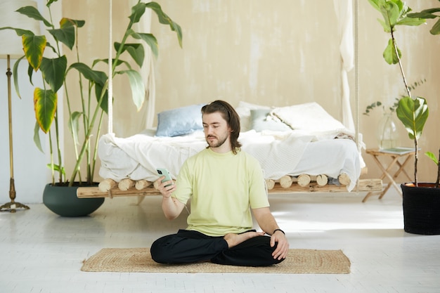 Hübscher Mann, der auf Yogamatte sitzt mit Telefon, Yogalehrer zu Hause hält Telefon für Online-Lektion