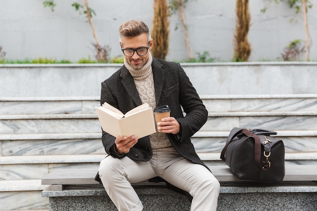 Hübscher Mann, der Jacke trägt, die ein Buch liest, während draußen sitzt