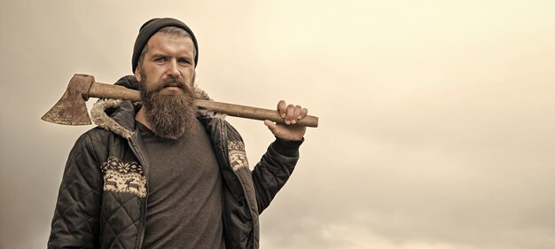 Hübscher Mann Hipster oder Kerl mit Bart und Schnurrbart auf ernstem Gesicht in Hut und Jacke hält rostige Axt mit Holzgriff im Freien vor bewölktem Himmel auf natürlichem Hintergrund, Kopierraum