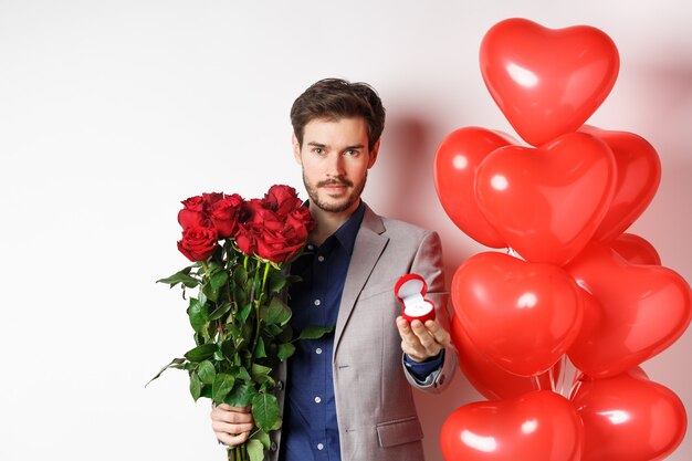 Hübscher Mann im Anzug, der einen Verlobungsring und einen Strauß roter Rosen gibt, heiraten mich am Valentinstag, stehend mit Herzballons auf weißem Hintergrund.