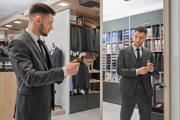 Hübscher Mann im eleganten gestreiften Anzug stehend, Spiegel in der Boutique betrachtend und Kleidung wählend.