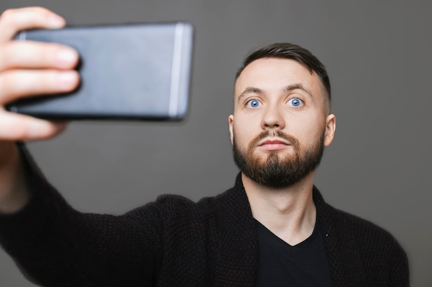 Hübscher Mann im stilvollen Outfit, das Smartphone hält und Kamera betrachtet, während für Selfie auf grauem Hintergrund aufwirft