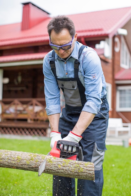 Hübscher Mann in der Arbeitsuniform, die Holz mit elektrischer Säge nahe Haus sägt