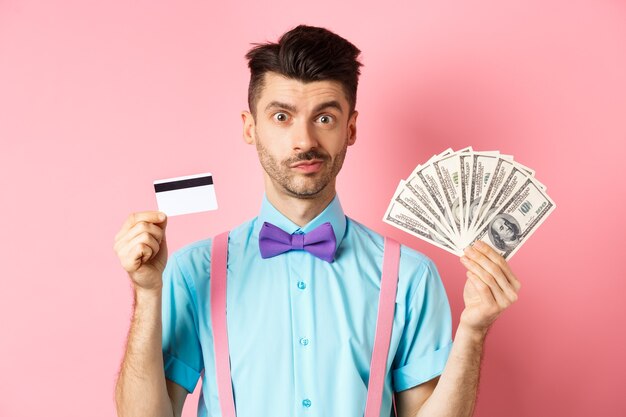Hübscher Mann in der Fliege, die Plastikkreditkarte und Geld in Dollar zeigt, ein Angebot machend, über rosa stehend. Speicherplatz kopieren