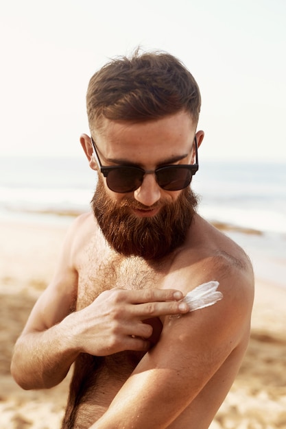 Hübscher Mann mit Bart, in Sonnenbrille Sonnenbaden mit Sonnenschutzlotion Körper im Sommer.