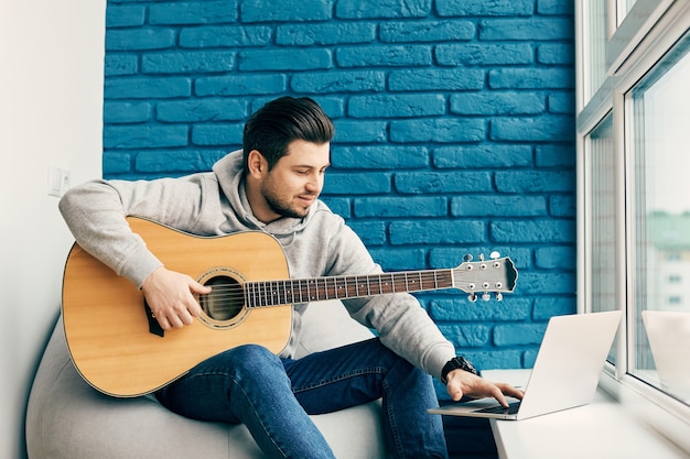 Hübscher Mann mit Gitarre, der seinen Laptop zu Hause mit der Wand aus blauen Ziegeln benutzt, Musiker, der übt, zu Hause zu palyen
