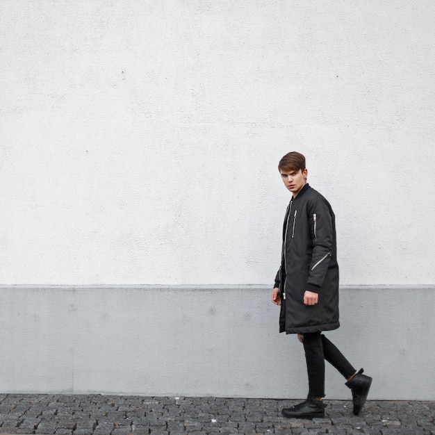 Hübscher Model-Typ in einer schwarzen Jacke und zerrissenen Jeans mit Turnschuhen, die nahe der Wand gehen