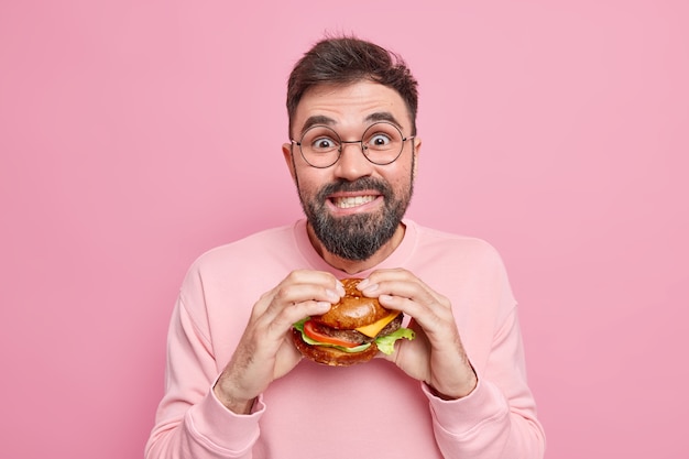 Hübscher positiver Kerl hat Essattacken ungesunde Ernährung hält appetitanregende Hamburger schaut gerne in die Kamera, die verlockend ist, Junk-Food zu essen, trägt runde Brillen