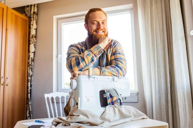 Hübscher rothaariger Mann mit langem Bart näht im Heimstudio an einer Nähmaschine