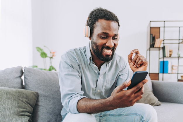 Hübscher schwarzer erwachsener Mann zu Hause, der online Musik mit Smartphone über Kopfhörer hört