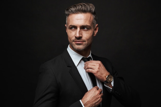 Hübscher selbstbewusster Geschäftsmann im Anzug, der isoliert über schwarzer Wand steht und posiert