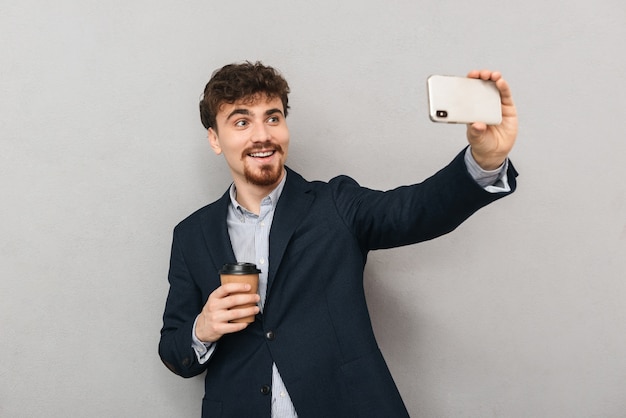 Hübscher selbstbewusster junger Geschäftsmann, der eine Jacke trägt, die isoliert über grau steht, ein Selfie nimmt und eine Tasse Kaffee zum Mitnehmen hält