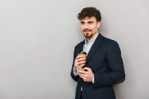 Hübscher selbstbewusster junger Geschäftsmann, der eine Jacke trägt, die isoliert über grau steht, unter Verwendung des Mobiltelefons, das Tasse Kaffee zum Mitnehmen hält