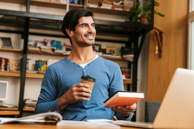 Hübscher selbstbewusster junger Mann, der am Bibliotheksschreibtisch sitzt, arbeitet / studiert, Laptop benutzt, Tasse Tasse Kaffee zum Mitnehmen hält