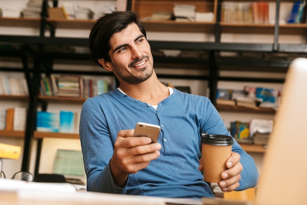 Hübscher selbstbewusster junger Mann, der am Bibliotheksschreibtisch sitzt, arbeitet / studiert, Laptop-Computer benutzt, Handy hält, Kaffee trinkt