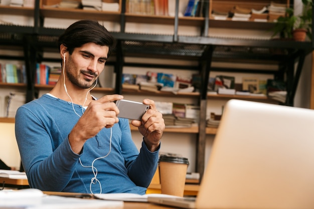 Hübscher sicherer junger Mann, der Kopfhörer trägt, die am Bibliotheksschreibtisch sitzen, arbeiten / studieren, Handy halten