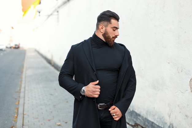 Hübscher starker Mann mit einem Bart in einem schwarzen trendigen Mantel geht auf der Straße