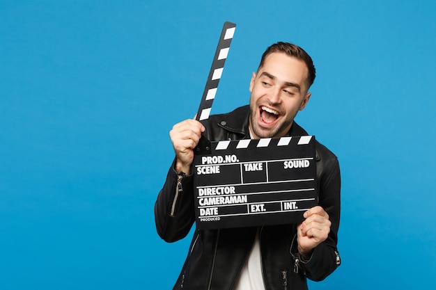 Hübscher, stilvoller junger unrasierter Mann in schwarzer Jacke, weißem T-Shirt, der in der Hand hält, um Filmklappe zu machen, die auf blauem Wandhintergrund Studioporträt isoliert ist. Menschen Lifestyle-Konzept. Kopieren Sie Platz.