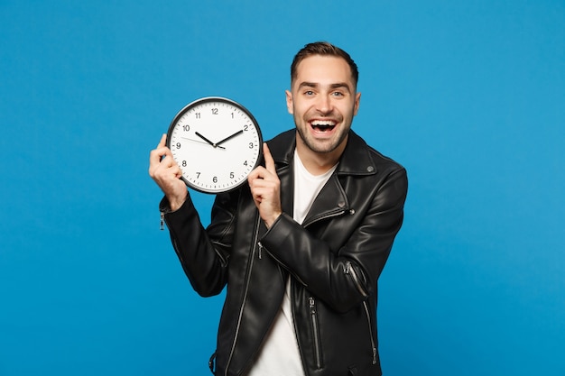 Hübscher, stilvoller junger unrasierter Mann in schwarzer Lederjacke, weißem T-Shirt, das eine runde Uhr hält, die auf blauem Hintergrundstudioporträt isoliert ist. Menschen Lifestyle-Konzept. Beeile dich. Kopieren Sie Platz.