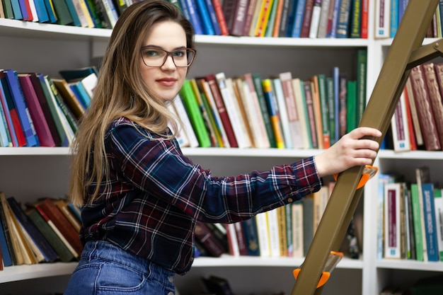 Hübscher Student steht auf einer Trittleiter in der Bibliothek, um das richtige Buch zu finden.