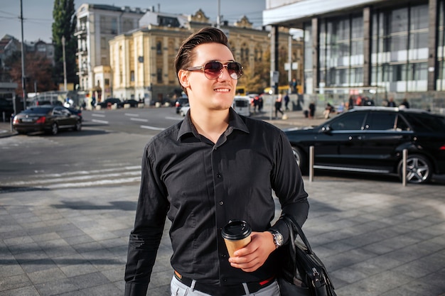 Hübscher und glücklicher junger Mann geht auf Straße. Er schaut durch die Sonnenbrille nach rechts. Junger Mann hat eine Tasse Getränk und Ledertasche. Er ist selbstbewusst.