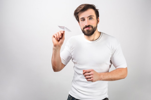 Hübscher und glücklicher Mann im weißen Hemd steht nahe der Wand und hält ein Papierflugzeug. Er ist bereit, es zu starten. Isoliert auf weiße wand.