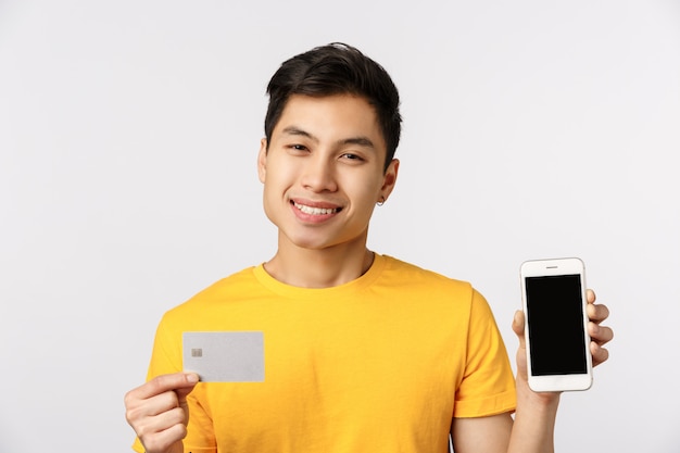 Hübscher und niedlicher lächelnder asiatischer Mann im gelben T-Shirt, zeigt Telefonanzeige und Kreditkarte, grinsend erfreut, empfehlen Bankdienst, Online-Zahlungssystem, stehende weiße Wand