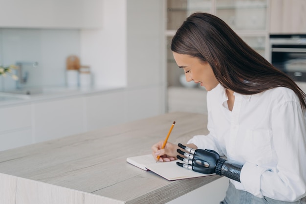 Hübsches behindertes Mädchen mit bionischer Armprothese schreibt in Notizbuch, das zu Hause am Küchentisch sitzt