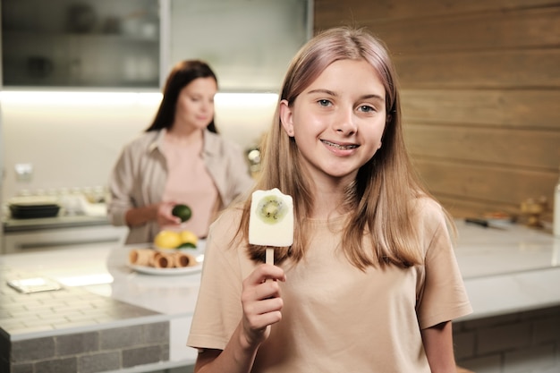 Hübsches blondes junges Mädchen, das Ihnen hausgemachtes Eskimo-Eis mit Scheibe frischer Kiwi zeigt, während es vor der Kamera in der Küche isst