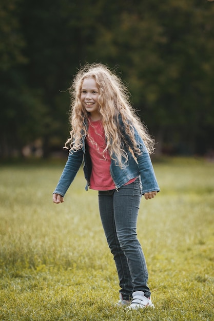 Hübsches blondes lächelndes Kind, das Jeansjacke im Park trägt, Porträt im Freien