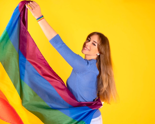 Hübsches blondes lesbisches Mädchen mit schwuler Stolzflagge auf pastellgelber Wand