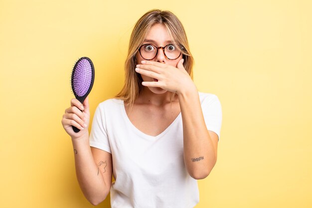 Hübsches blondes Mädchen, das den Mund mit den Händen mit einem schockierten bedeckt. Haarbürstenkonzept